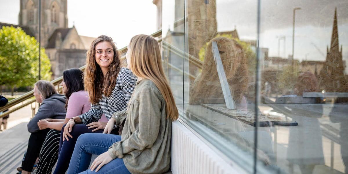 Two students sit on the Parkinson steps chatting. You can see a tree and the Emmanuel Centre in the background.