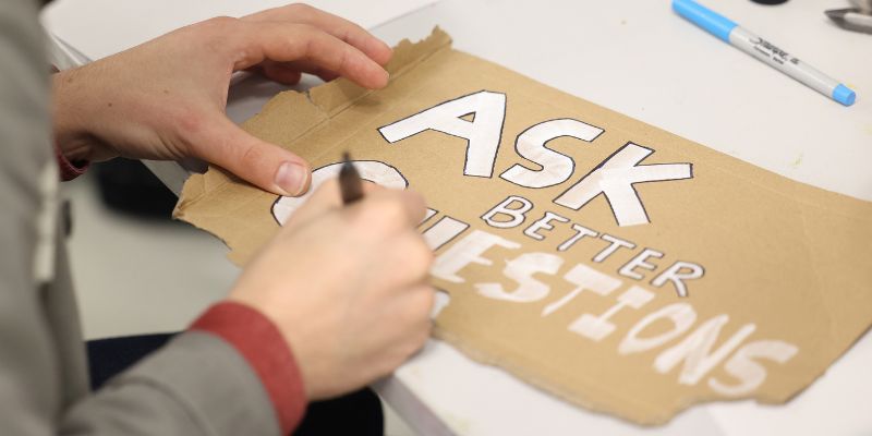 A piece of art, done on cardboard, with 'Ask better questions' written in white pen.