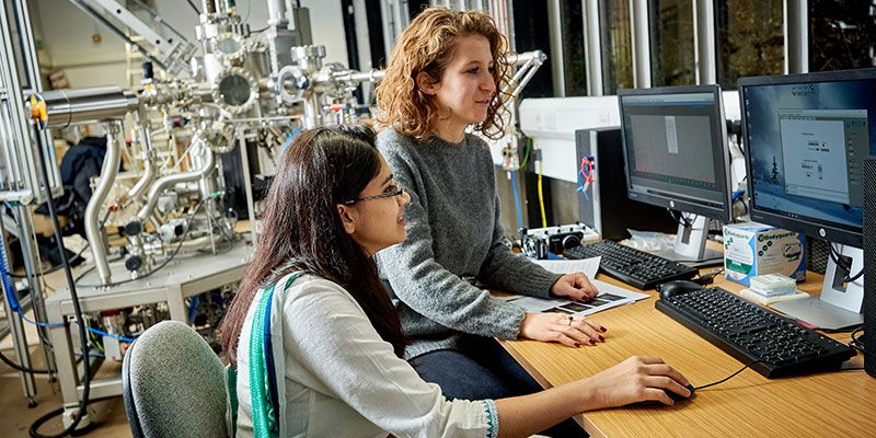Two researchers work on computers with a large piece of Bragg Centre equipment near their desk.