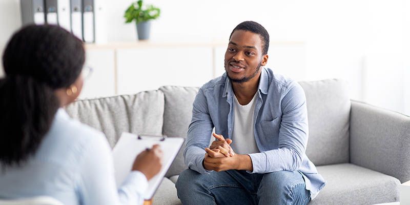 A man sat on a sofa talking to a clinician who is making notes on a clip board.