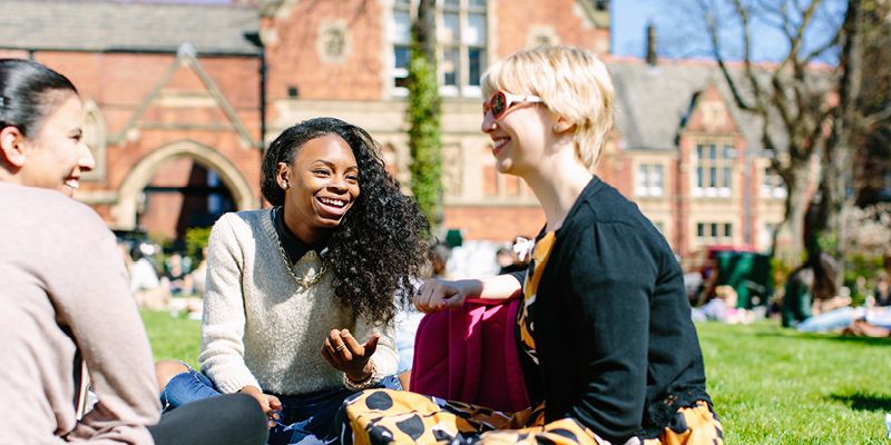 Three students sit on a patch of grass on campus, with a red brick building in the background.