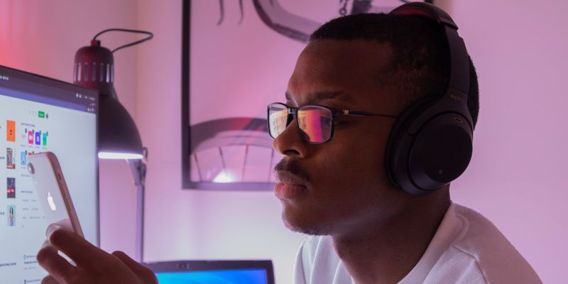 A person wearing headphones, looking at a mobile phone, in front of computer screens.