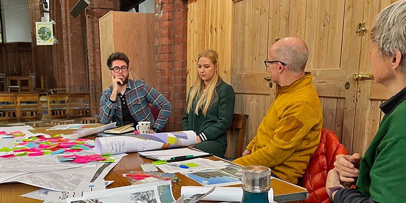 A group of people sat around a table with paper plans, contributing to community-led urban planning.