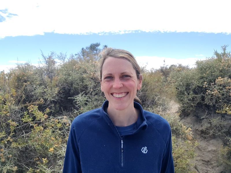 A woman smiling at the camera with shrubs behind her and a blue sky.