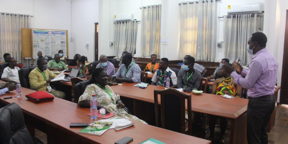 A classroom full of people in Tanzania, sat in rows of desks, in the middle of a lively discussion with someone stood up at one end of the room.