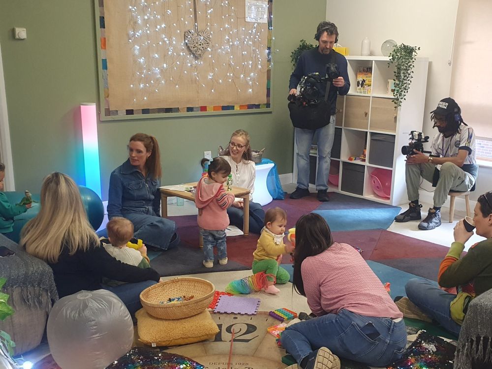 A group of parents and young children sitting on the floor, talking to researchers about baby food products while being filmed