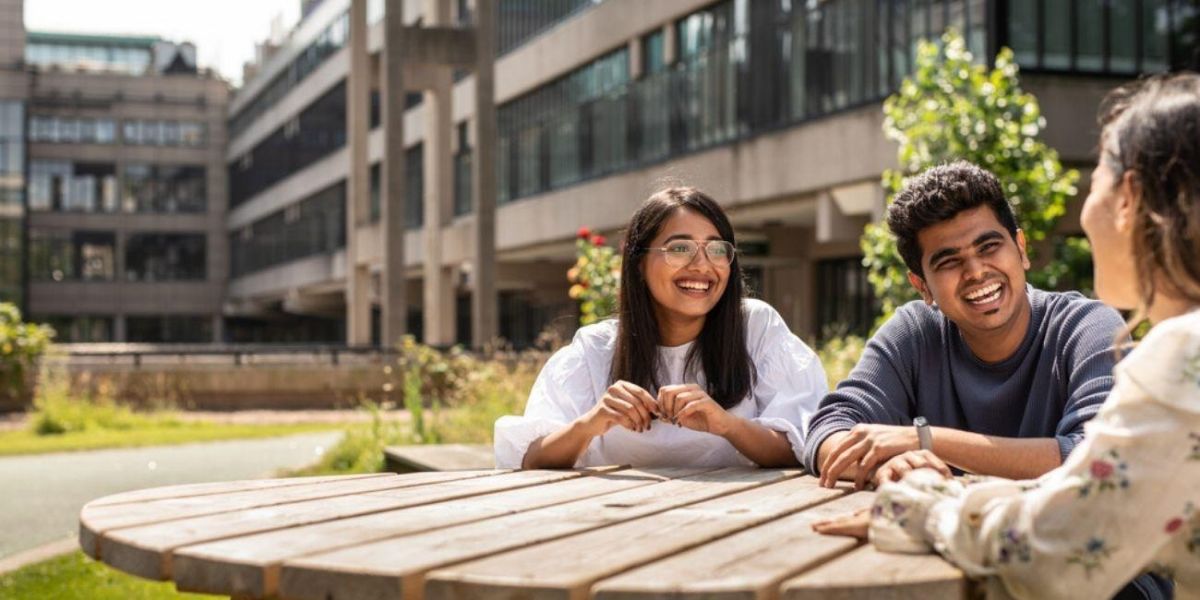 Three laughing students sit around a picnic table on campus.