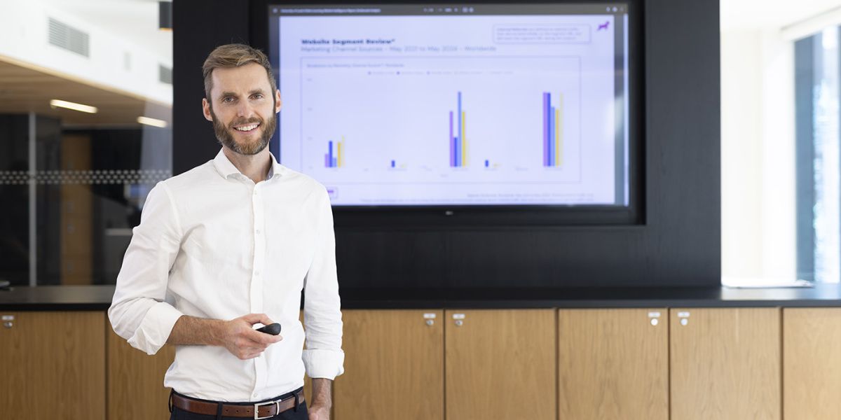 A student in shirt and trousers stood in front of a screen displaying bar charts and data.