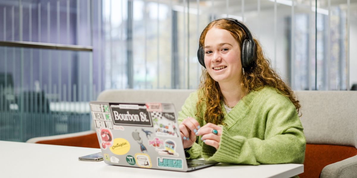 A smiling student, wearing headphones, sits at a table with a laptop.