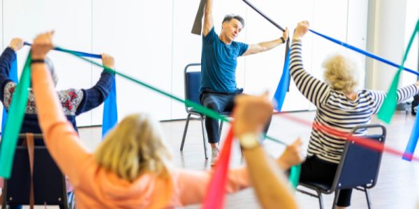 Older people take part in an exercise class on chairs, holding colourful resistance bands over their heads to stretch their arms.