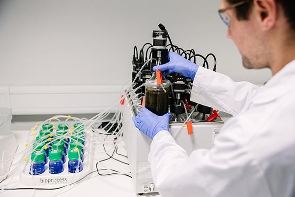 A person in a lab coat and gloves using a machine that is linked to bottles containing liquid