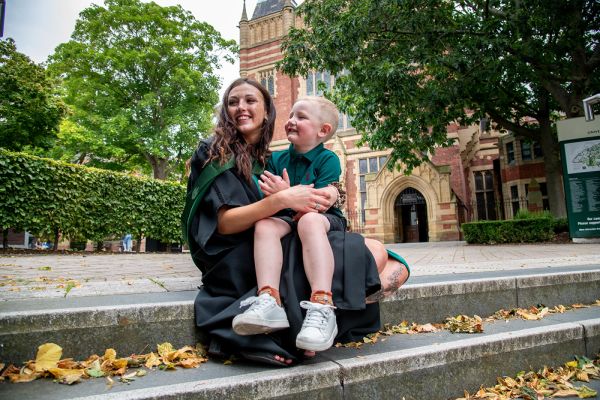 Emilie Barron, wearing a graduation gown, sitting on a step outside the Great Hall with a small child on their knee.