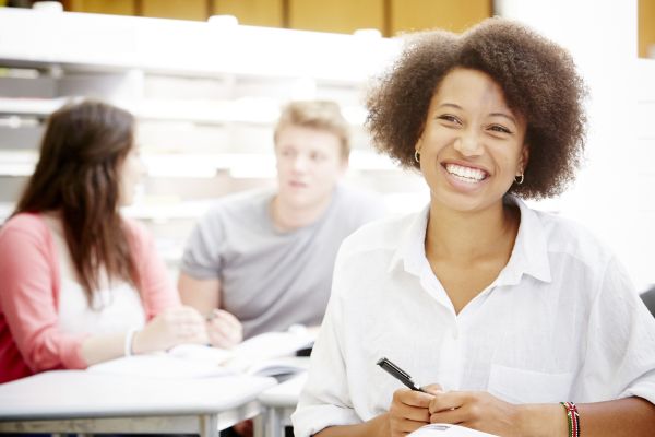 A student sits at a desk, holding a pen, smiling. Two students talk in the background.