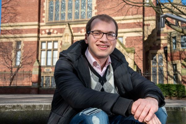Makeen Baroudi, smiling, sitting on a step in front of the Great Hall.