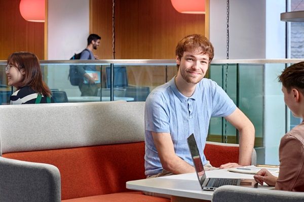 A student sat in a modern study space smiling at the camera. other students are working on laptops or chatting