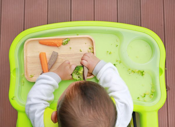 A baby sitting at a highchair with vegetable sticks on the tray in front of them, seen from above