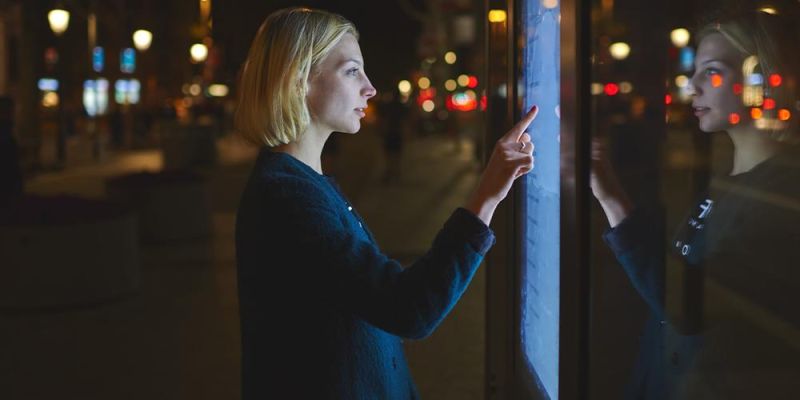 A person stands in a dark street at night pressing a touch screen on a board.