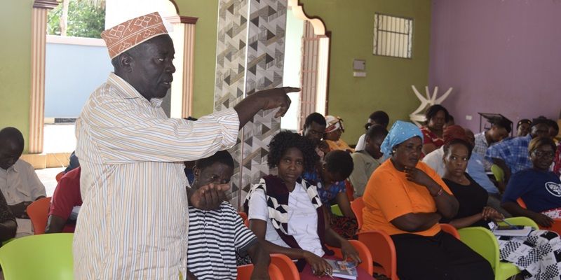 A room full of people in Tanzania taking part in a lively discussion. One person is stood up, speaking and gesturing