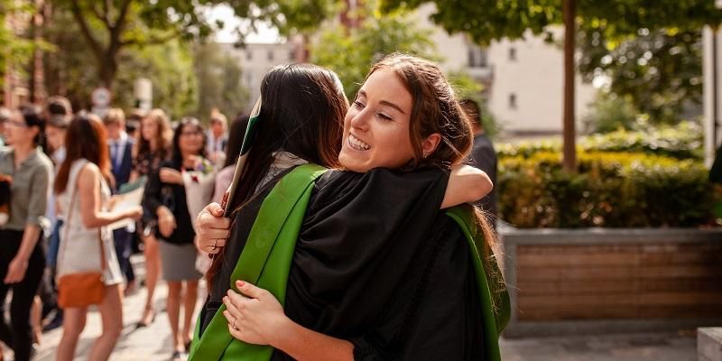 Two students wearing graduation gowns and hugging outside the Great  Hall