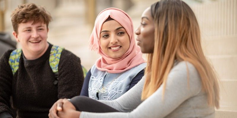 Three students of different ethnicities are sitting on the steps of the University's Parkinson Building talking.