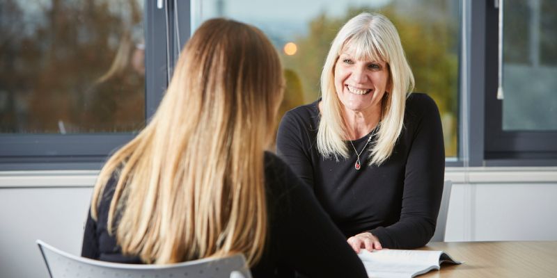 One person sat at a desk with a book in front of them talking to another person whose face is not shown.