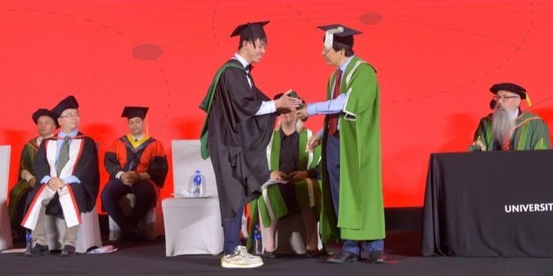  A student stands on stage in their graduation robes, receiving a scroll from Professor Hai-Sui Yu, Provost and Deputy Vice-Chancellor of the University of Leeds, as other academics, also in graduation attire, sit on stage, watching.