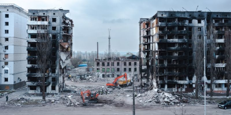 Fire-blackened multi-storey buildings destroyed by war in Ukraine, stand in rubble, beside diggers.