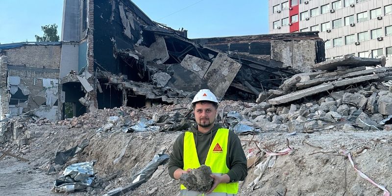 Engineer wearing a hi-vis jacket and holding a piece of rubble, stands in front of the remains of a building in Ukraine that has been destroyed during the war.