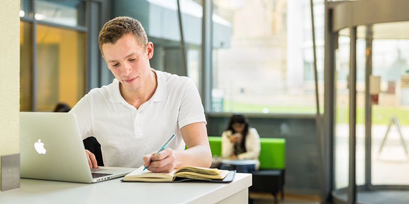 An adult sits in a café with a laptop, writing notes in a notebook.