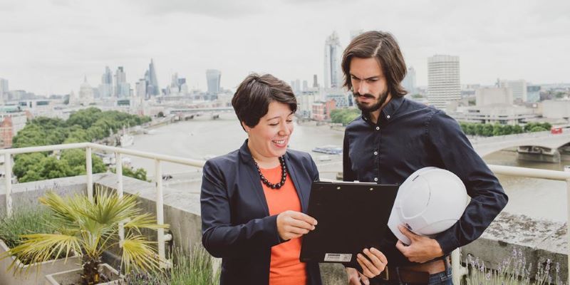 Two individuals stood on a balcony with a cityscape behind them, looking at a computer tablet.