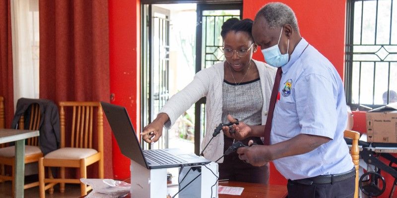 Two people standing looking at a laptop screen. One of them, Miss Johnny Odu, is talking while pointing at the screen. The other person is holding keyhole surgery devices in each hand.