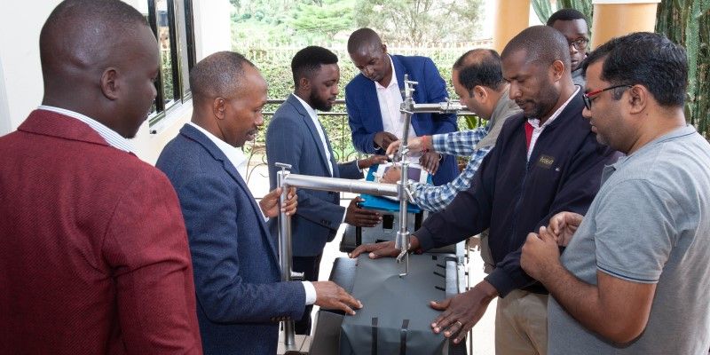 A group of people standing around a table, interacting with metal devices for keyhole surgery. The device in view has a spiral arm lifting up the top of a container.